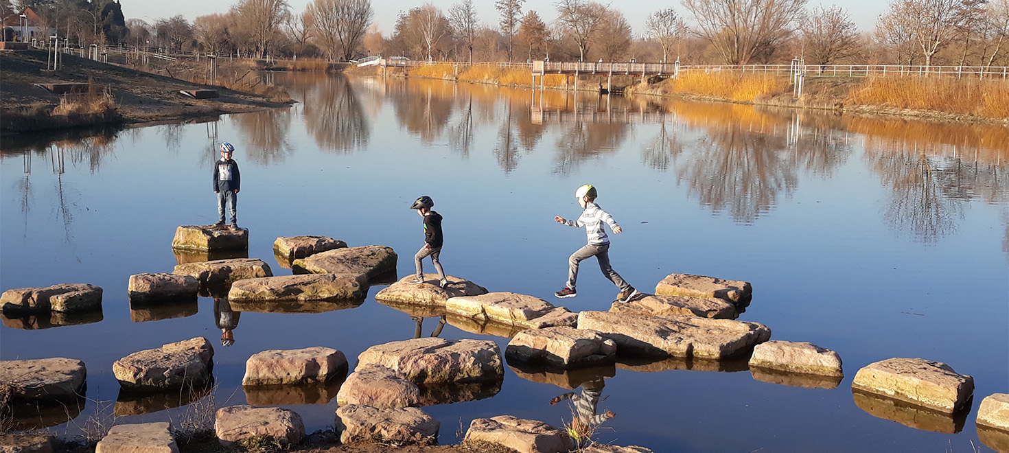 Kinder überqueren Steine an einem renaturierten Flussufer, im Hintergrund fährt ein Frachtschiff auf dem Neckar